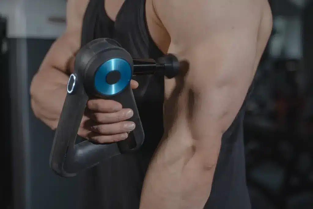Um homem musculoso usando um massageador elétrico preto e azul em seu bíceps, demonstrando recuperação muscular pós-treino ou alívio da dor, com fundo desfocado de academia.