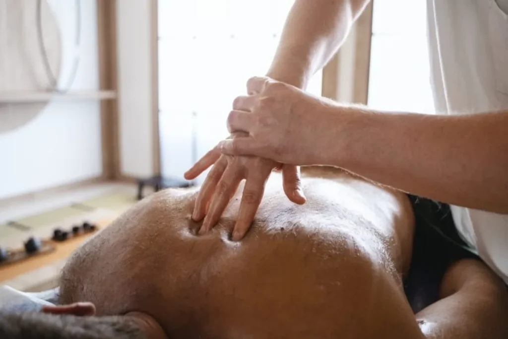 Um homem deitado de bruços em uma maca, recebendo uma massagem nas costas. As mãos do terapeuta aplicam pressão e óleo, simbolizando a prática de um terapeuta tantra.