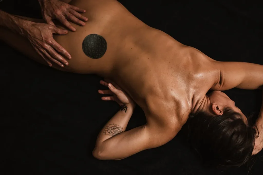 Uma pessoa deitada de bruços em uma maca escura, recebendo uma massagem relaxante na região lombar. A pele está oleada e há uma tatuagem de geometria sagrada na parte inferior das costas, simbolizando a abordagem holística do tantra.