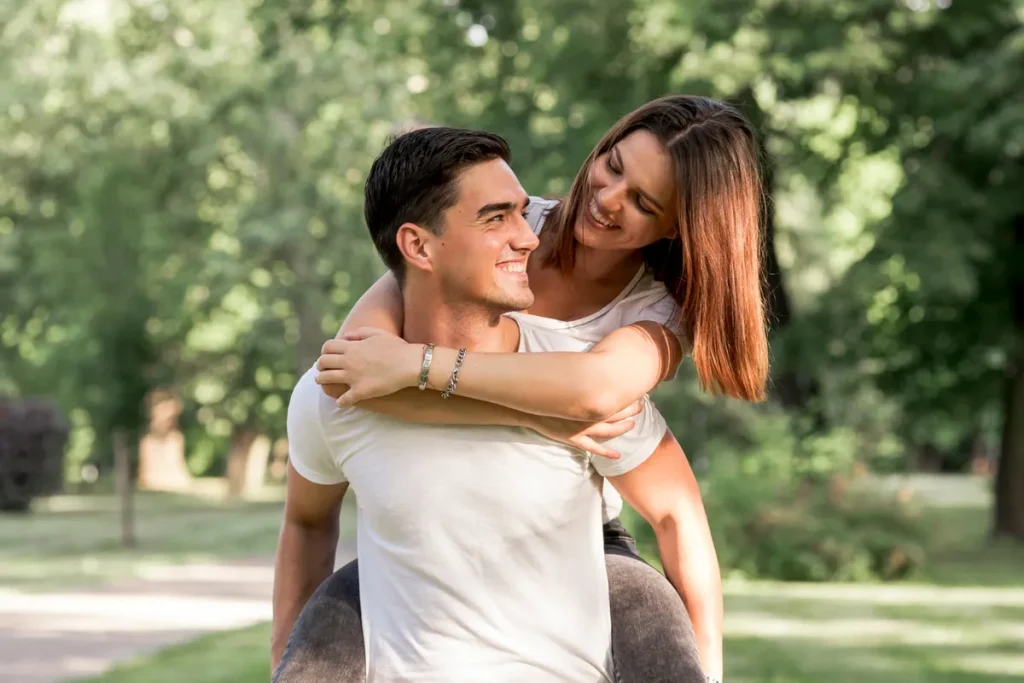 Casal sorridente e feliz, com a mulher nas costas do homem em um parque, representando a alegria e a "libido" no relacionamento.