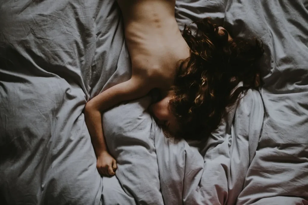 Uma foto com iluminação suave (low key) de uma pessoa deitada de barriga para cima em uma cama com lençóis cinzas amassados. A pessoa está sem camisa, com os braços relaxados e o cabelo castanho espalhado pelo travesseiro. A postura de exaustão e relaxamento profundo após a intimidade simboliza o momento que sucede o clímax ou a ejaculação, focado no descanso e no bem-estar pós-sexual.