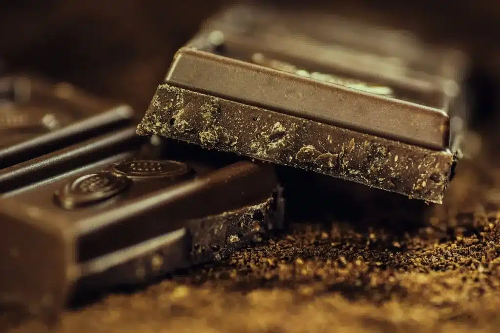 Barras de chocolate escuro quebradas e espalhadas sobre cacau em pó ou terra, com um foco macro que realça a textura rugosa do chocolate.