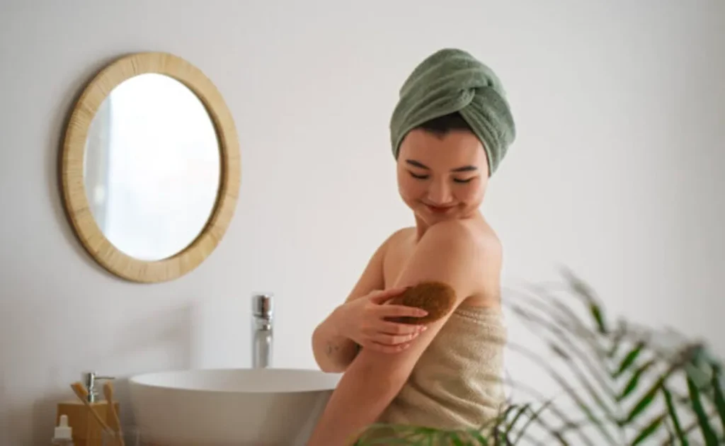Uma mulher envolta em uma toalha bege e com uma toalha verde enrolada na cabeça está sorrindo enquanto esfolia suavemente o braço com uma escova ou luva esfoliante natural. O fundo é um banheiro claro com uma pia e um espelho redondo de moldura de bambu, simbolizando um momento de autocuidado, relaxamento e atenção à saúde da pele.