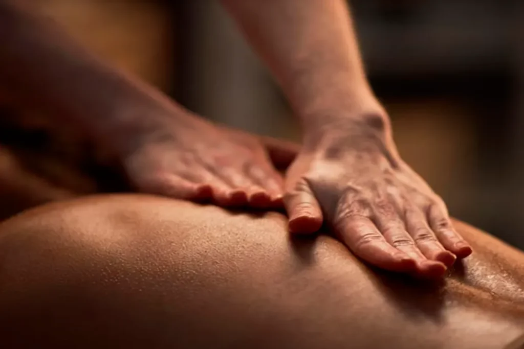 Close-up das mãos de um terapeuta aplicando pressão e deslizando sobre as costas de um paciente, que está deitado de bruços. O toque suave e a iluminação quente focam nos músculos e na coluna vertebral, ilustrando uma sessão de massagem relaxante ou terapêutica conhecida em alguns contextos como massagem da donzela.