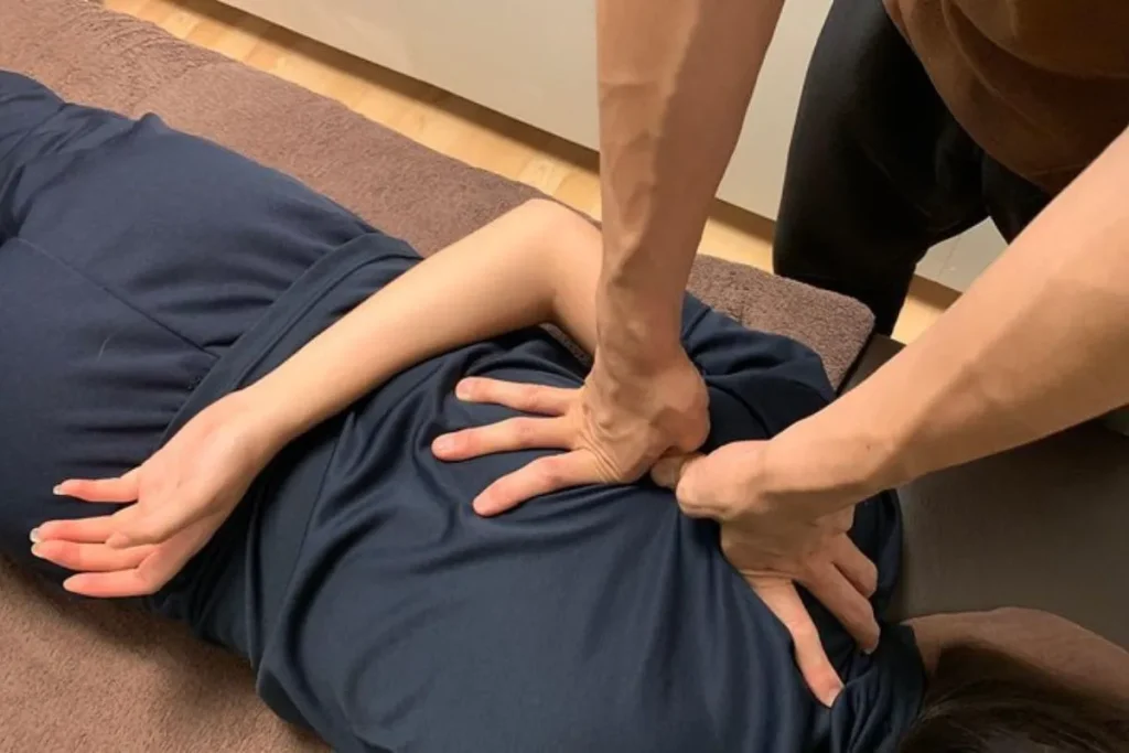 Close-up das mãos fortes de um terapeuta aplicando pressão intensa nas costas de um paciente, que está deitado de bruços, vestindo roupas escuras. O foco está na técnica de massagem japonesa Shiatsu ou Anma, utilizada para liberar tensões musculares.