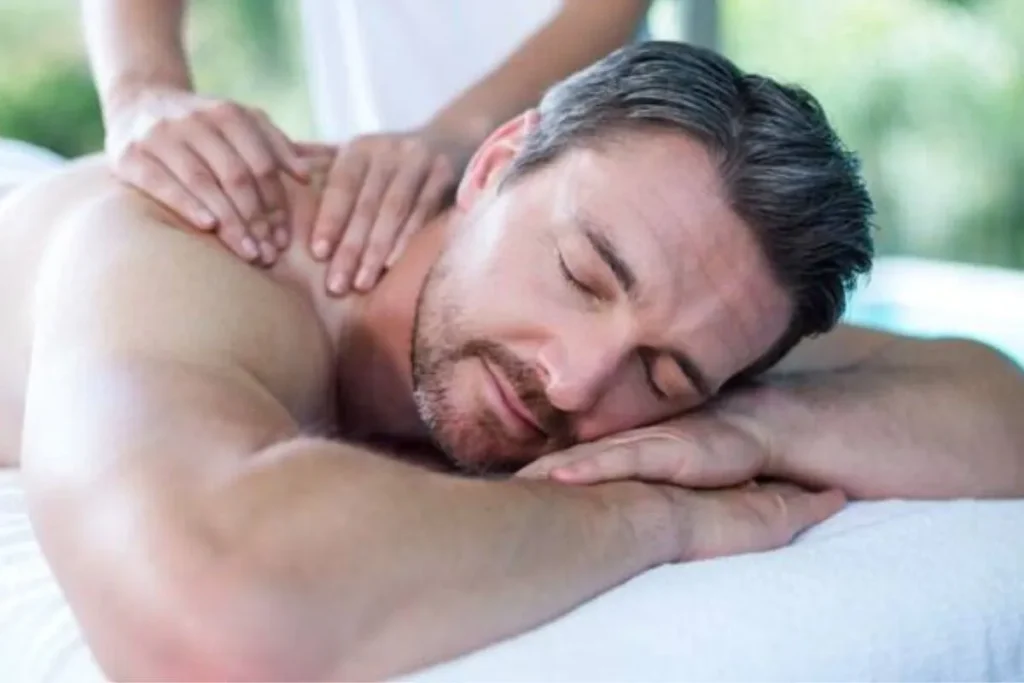 Close-up de um homem adulto deitado de bruços em uma maca, com os olhos fechados e uma expressão de paz, recebendo uma massagem relaxante corpo inteiro nas costas de uma terapeuta. A cena transmite tranquilidade e conforto.