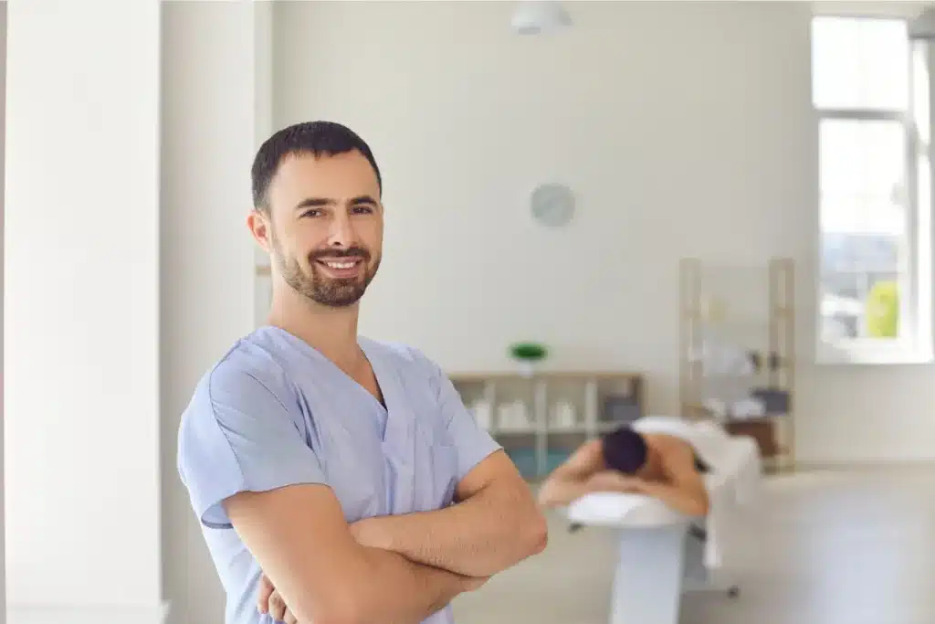 Massagista profissional com uniforme azul sorrindo para a câmera em uma clínica de massagens, com um cliente relaxando ao fundo.