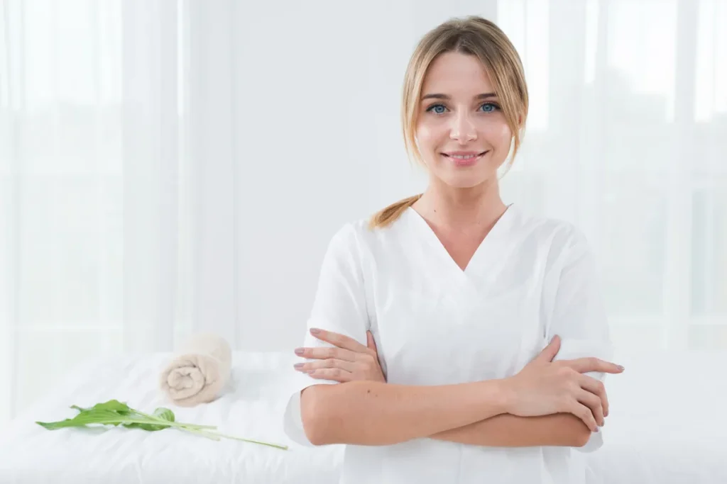 Mulher massoterapeuta sorrindo, em uniforme branco, com uma maca de massagem ao fundo.