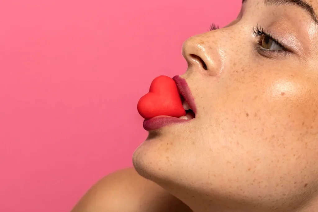 Close no rosto de mulher com batom rosa segurando um pequeno coração vermelho entre os lábios, sobre fundo rosa