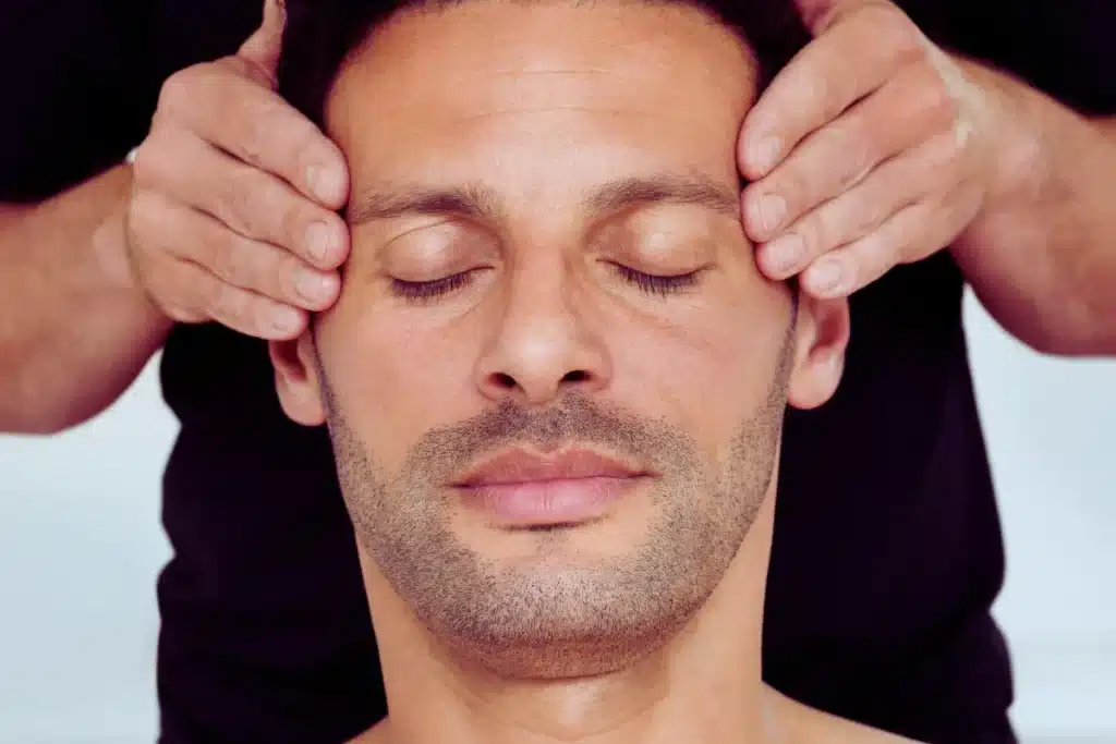Homem com expressão serena recebe uma massagem terapêutica na cabeça, com as mãos do massagista aplicando leve pressão nas têmporas.