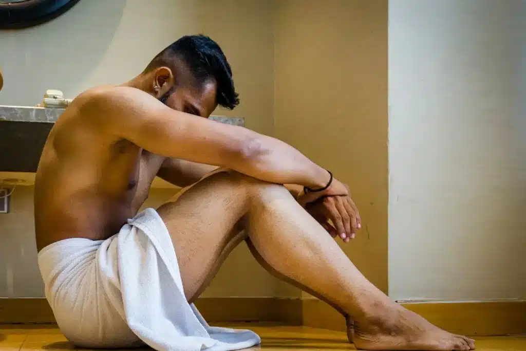 Homem de toalha branca sentado no chão, com a cabeça baixa e os braços sobre os joelhos, em um ambiente interno que parece ser um banheiro ou quarto. massagem peniana