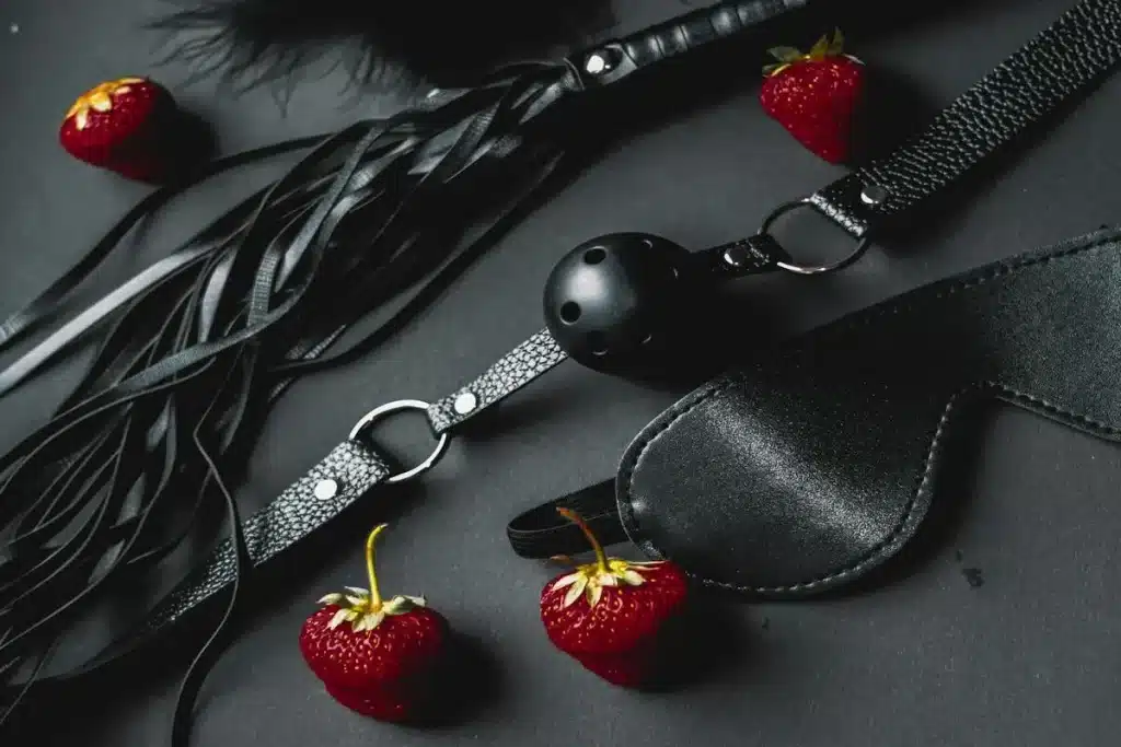 Acessórios BDSM como chicote, venda, mordaça e morangos sobre fundo escuro representando erotismo e fetiches