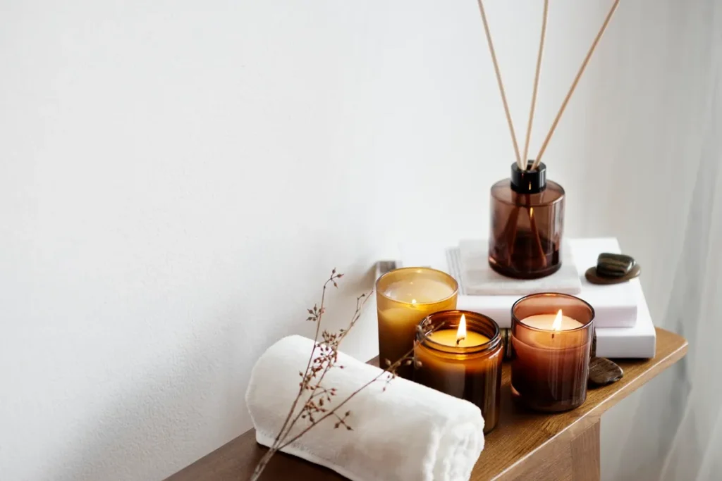 Velas aromáticas acesas, difusor de varetas, toalha branca e pedras sobre bancada de madeira, criando ambiente de aromaterapia