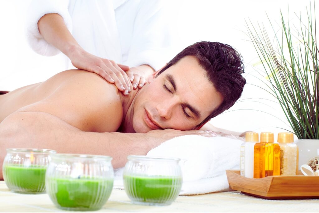 Homem deitado de bruços recebendo massagem nos ombros em ambiente de spa com velas e produtos de cuidado.
