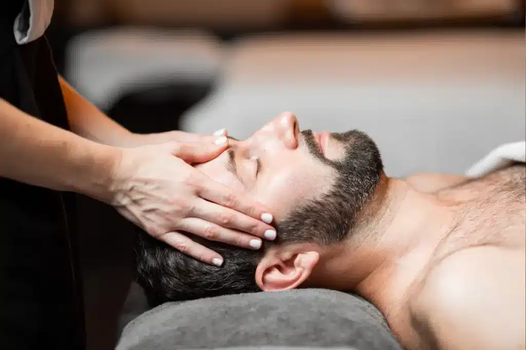 Homem deitado de costas, olhos fechados, recebendo massagem na cabeça por terapeuta em ambiente de spa