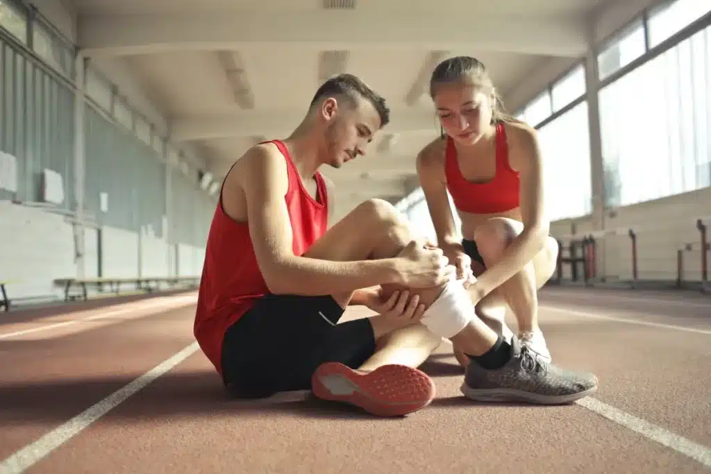 Uma mulher jovem em roupa esportiva vermelha ajuda um homem com cãibra ou lesão na perna, sentados em uma pista de corrida interna, aplicando uma bandagem no tornozelo dele.