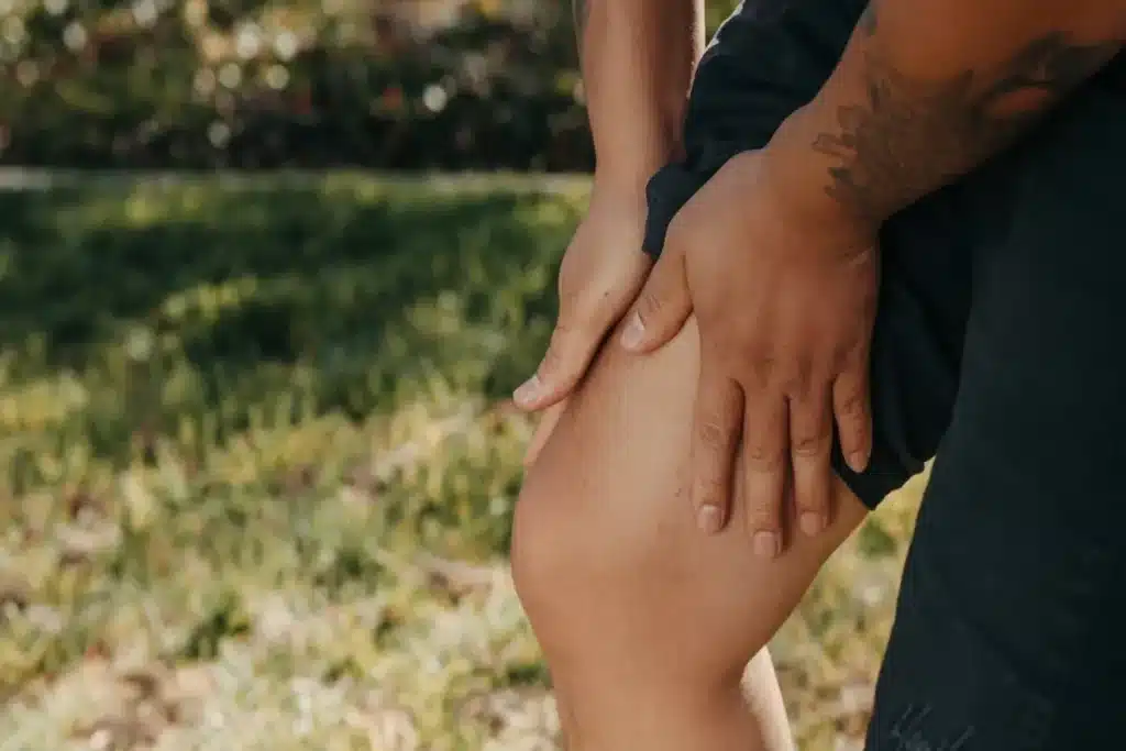 Close-up de uma pessoa com shorts escuros e braços tatuados segurando a coxa, indicando dor muscular ou cãibra, em um ambiente externo com gramado desfocado.