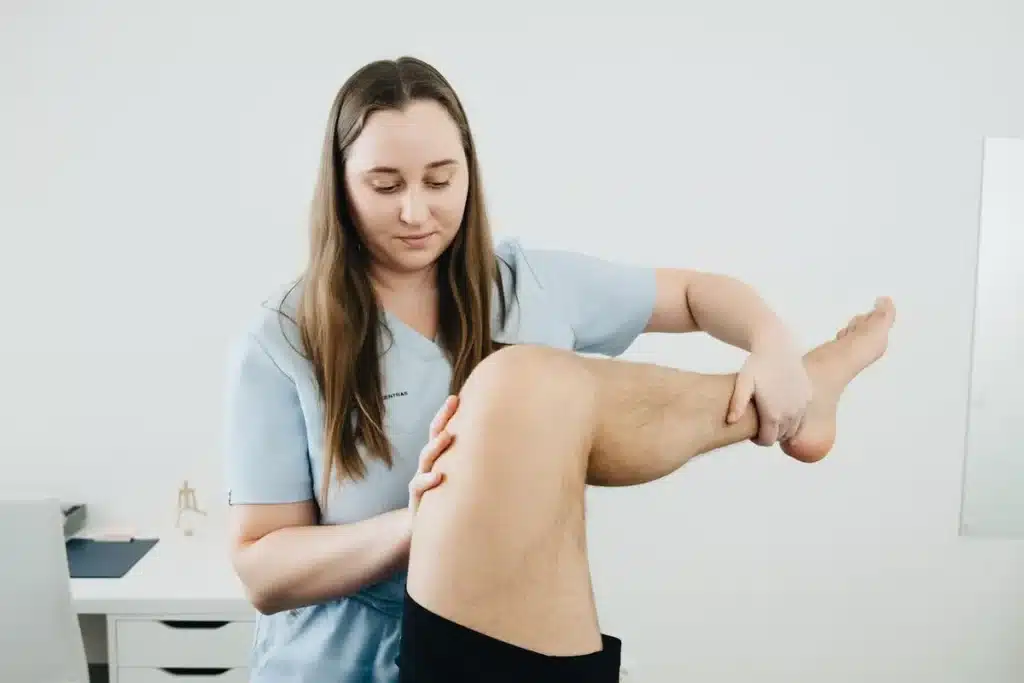 Fisioterapeuta feminina em uniforme azul claro examinando a perna de um paciente deitado, elevando e manipulando o joelho e a panturrilha, em uma clínica branca e iluminada.