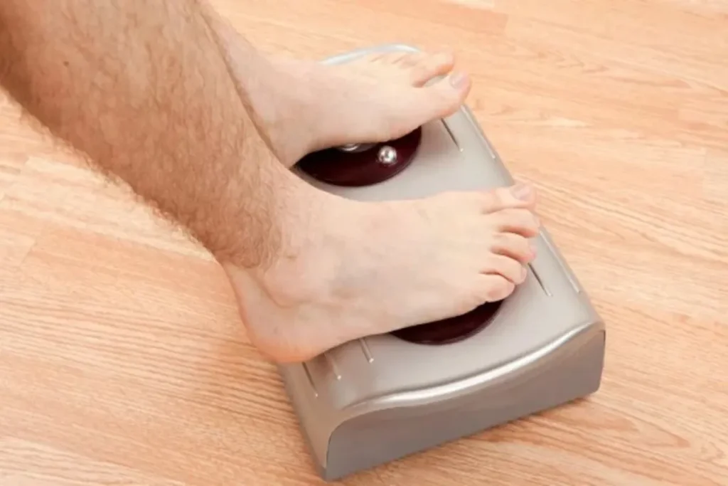 Pés de uma pessoa em um massageador elétrico, com a vibração ativada, em um piso de madeira.