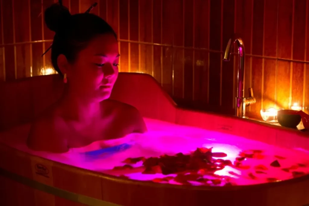 Mulher em banheira de madeira com água iluminada por luz vermelha (cromoterapia), pétalas de rosa flutuando e velas ao redor.