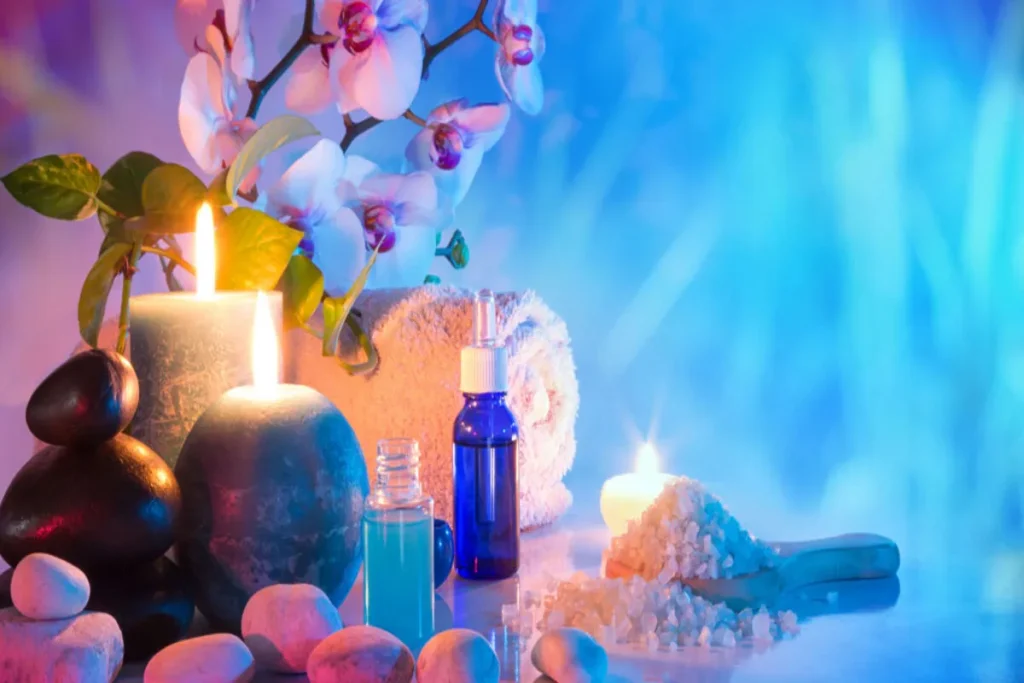 Itens de cromoterapia e spa com velas acesas, óleos essenciais em frascos azuis e sal de banho, com orquídeas e pedras em um ambiente de luz azul e rosa.