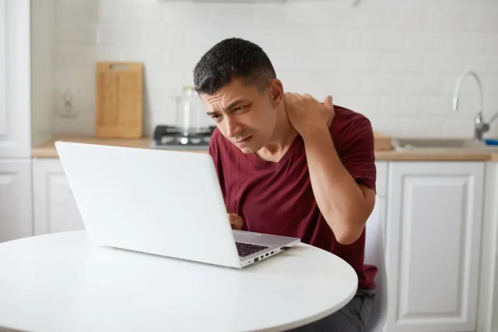 Homem com expressão de dor segurando o pescoço e o ombro enquanto trabalha em um laptop em uma mesa de cozinha, ilustrando má postura ergonômica.