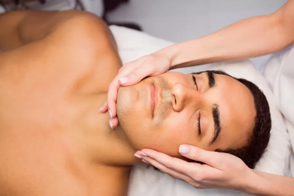 Um close-up de um homem de olhos fechados, deitado, recebendo uma massagem facial, com as mãos do terapeuta em seu rosto.