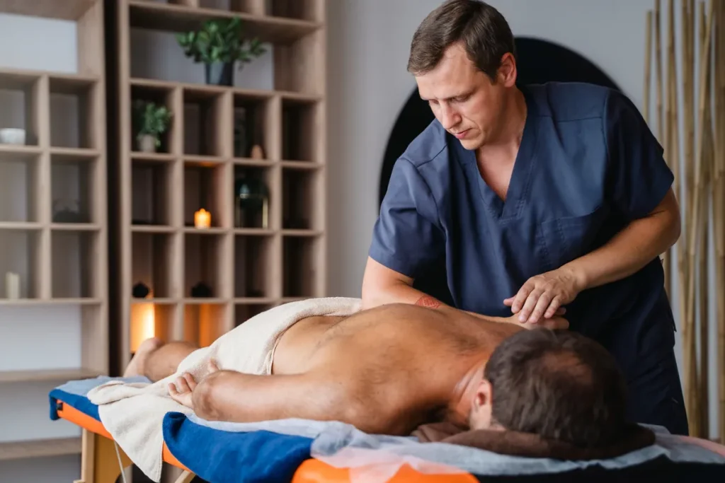 Um massagista em uniforme azul segura uma garrafa de gel enquanto realiza uma massagem nas costas de um cliente deitado em uma maca de tratamento.