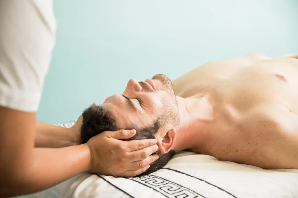 Homem deitado em maca recebendo massagem relaxante na cabeça e no pescoço de um terapeuta, técnica que estimula o relaxamento e a produção dos hormônios do sono (serotonina e melatonina).