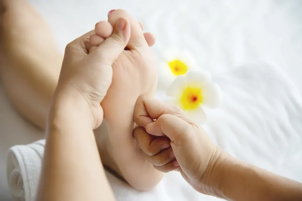 Duas mãos massageando a planta do pé de um cliente em uma maca, com duas flores brancas ao lado.
