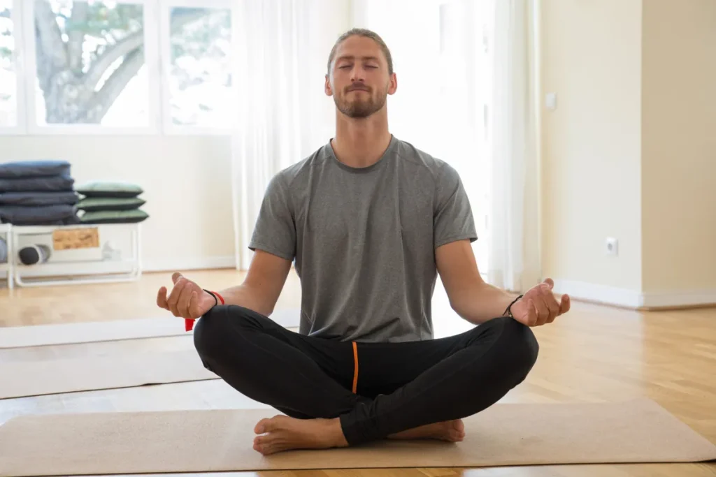 Homem caucasiano, com barba e cabelo comprido, praticando Tantra Yoga na postura fácil (Sukhasana) e meditando em um estúdio iluminado.