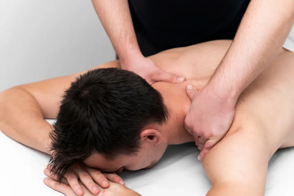 Um close-up das mãos de um massoterapeuta aplicando pressão e massagem no pescoço e ombros de um cliente masculino deitado de bruços.
