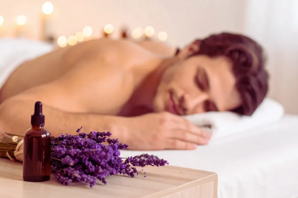 Homem deitado de bruços em maca de massagem, relaxando em ambiente com velas acesas, frasco de óleo essencial e flores de lavanda em primeiro plano.