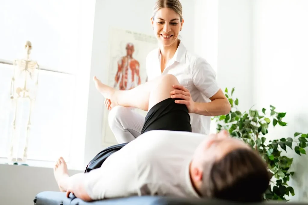 Fisioterapeuta mulher sorrindo e avaliando ou aplicando tratamento no quadril e joelho de um paciente deitado em uma maca. A cena, em um consultório claro, representa a reabilitação de dor pélvica.