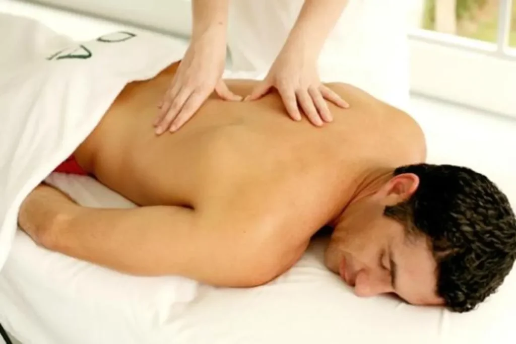 Vista de cima de um terapeuta aplicando massagem masculina nas costas de um homem deitado de bruços, em ambiente claro e relaxante.