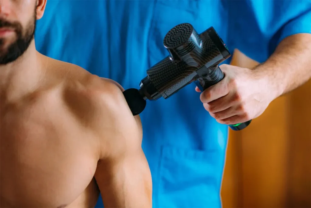 Terapeuta de azul aplicando uma pistola de massagem (dispositivo de percussão) no ombro musculoso de um cliente, para liberação miofascial.