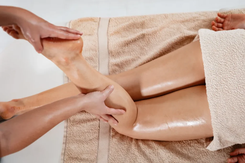 Mãos realizando massagem com óleo na perna de uma pessoa deitada com toalha cobrindo o corpo