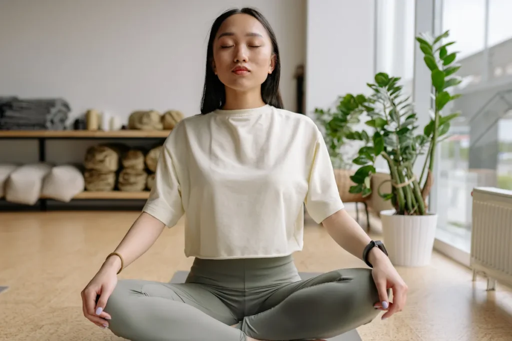 Mulher sentada em posição de meditação com olhos fechados praticando respiração consciente em ambiente interno com plantas.