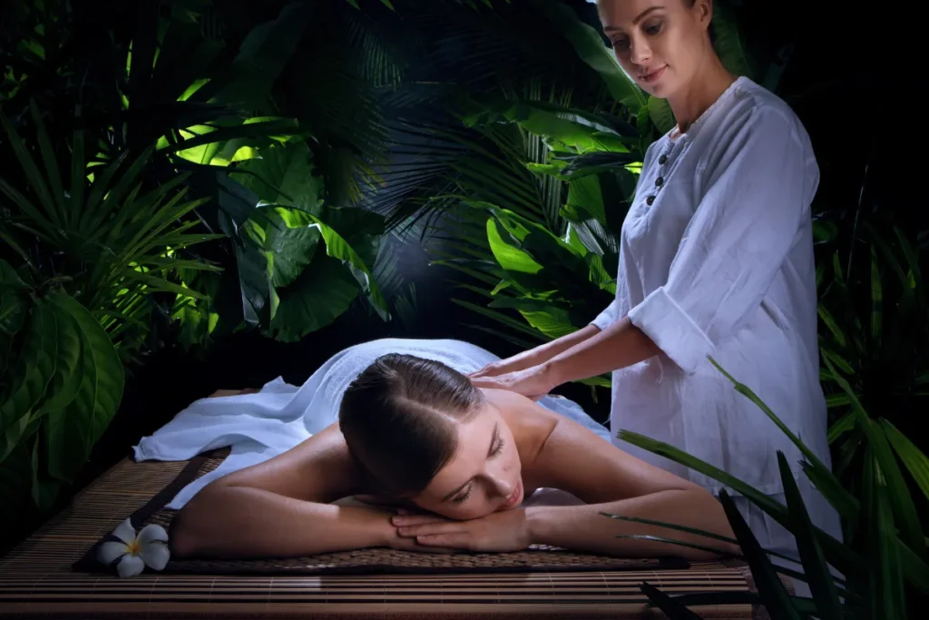 Mulher recebendo massagem nas costas enquanto está deitada em uma maca em ambiente de spa com plantas ao redor.