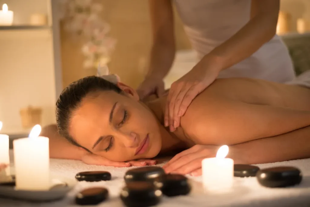 Mulher recebendo massagem relaxante nos ombros em spa com velas acesas e pedras de massagem sobre a mesa.