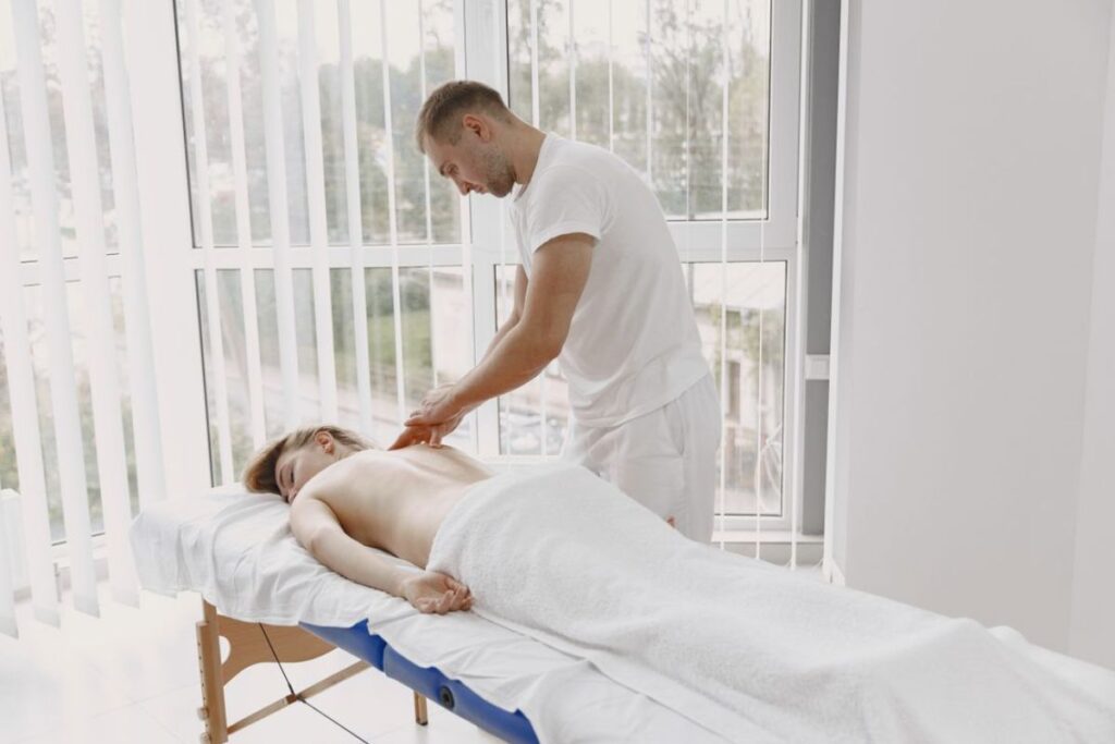 Profissional realizando massagem nas costas de uma cliente deitada em uma maca dentro de um consultório iluminado.