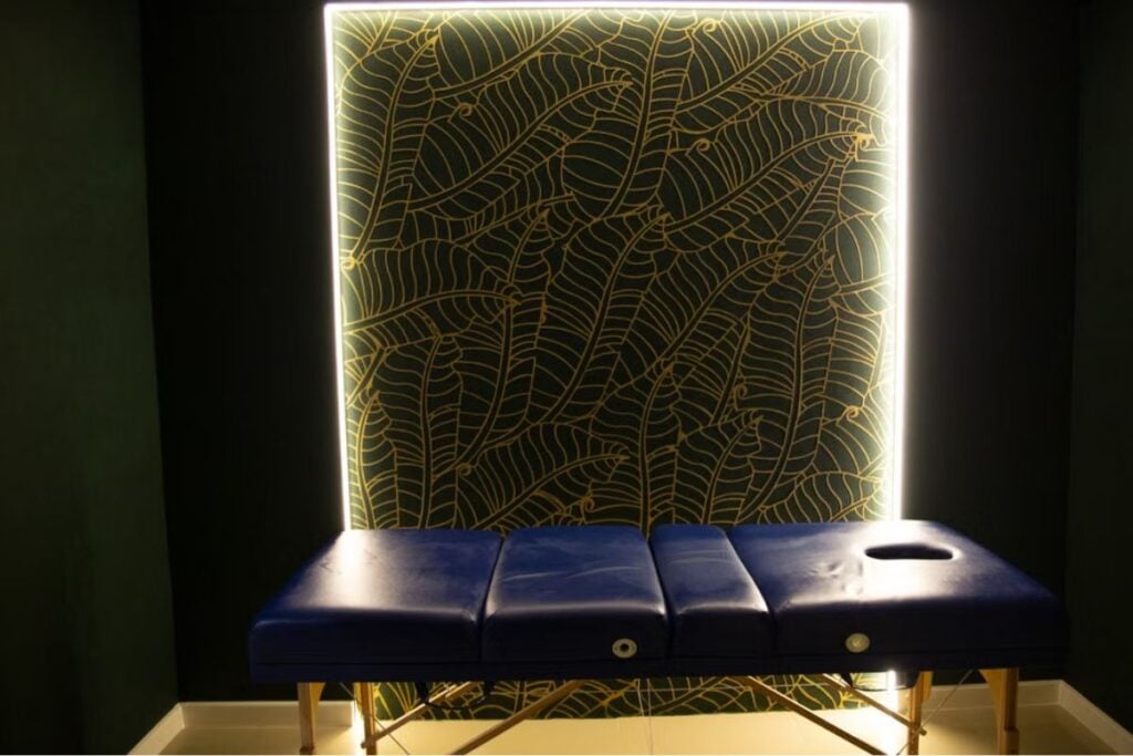Uma maca de massagem azul posicionada em frente a uma parede decorada com padrão de folhas e iluminação embutida.
