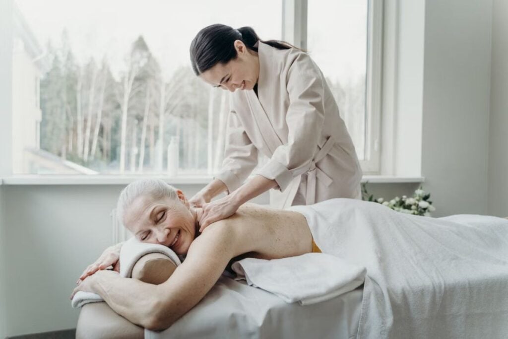 Uma profissional de massagem aplica técnica de massagem nas costas de uma mulher sorridente que está deitada em uma maca.