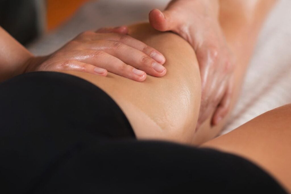 Mãos realizando massagem com óleo na região do quadril e glúteo durante sessão de recuperação muscular.