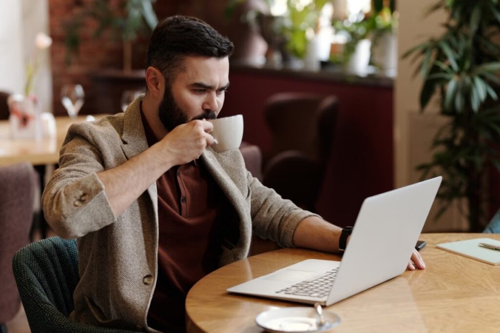 Fotografia de plano médio de um homem de barba e blazer bege sentado à mesa redonda de madeira, focado na tela de um notebook aberto enquanto toma uma bebida em uma xícara de cerâmica branca, em um ambiente interno de cafeteria.