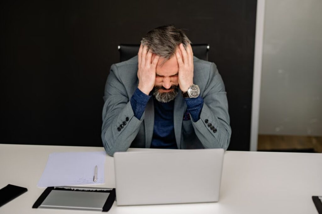 Fotografia de plano médio mostrando um homem de meia-idade, com barba grisalha e terno cinza, sentado à mesa de trabalho com as mãos nas têmporas e os olhos fechados em uma expressão de forte dor de cabeça ou estresse, diante de um notebook aberto.