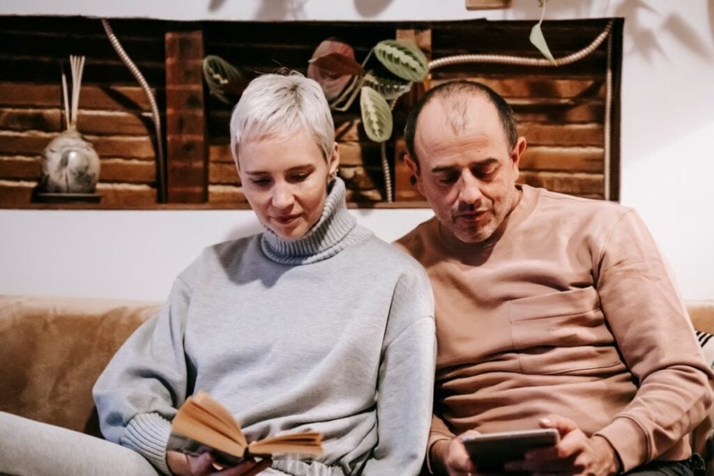 Fotografia de plano médio mostrando um homem calvo de blusa marrom-clara olhando para um tablet, enquanto uma mulher de cabelos loiros curtos e blusa de gola alta cinza lê um livro, sentados juntos em um sofá diante de uma parede decorada com madeira e plantas.