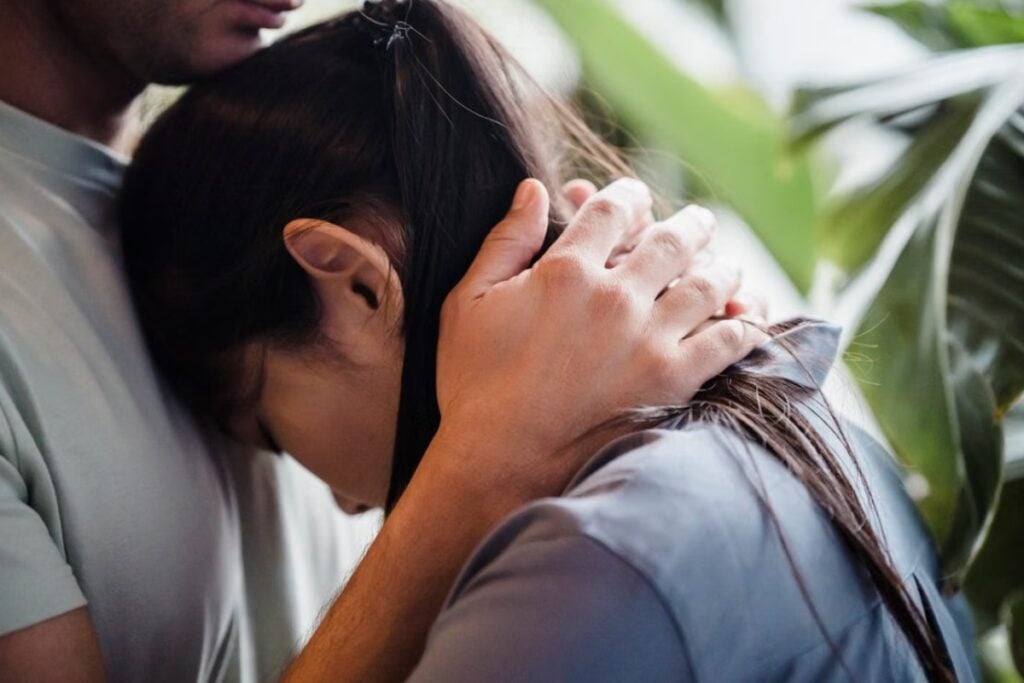 Fotografia em plano detalhe aproximado mostrando uma mulher de cabelos escuros com a cabeça apoiada no peito de seu parceiro, enquanto ele a envolve em um abraço protetor, acariciando sua nuca com a mão direita, cercados por folhas verdes ao fundo.