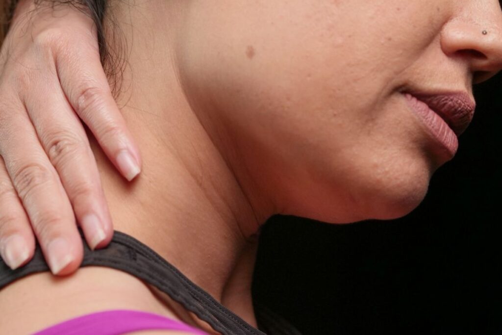 Fotografia em super close-up focando no perfil inferior do rosto, mandíbula e pescoço de uma mulher de pele morena, onde uma mão espalmada toca a lateral da região cervical sobre uma alça de top esportivo roxo e preto, contra um fundo escuro.