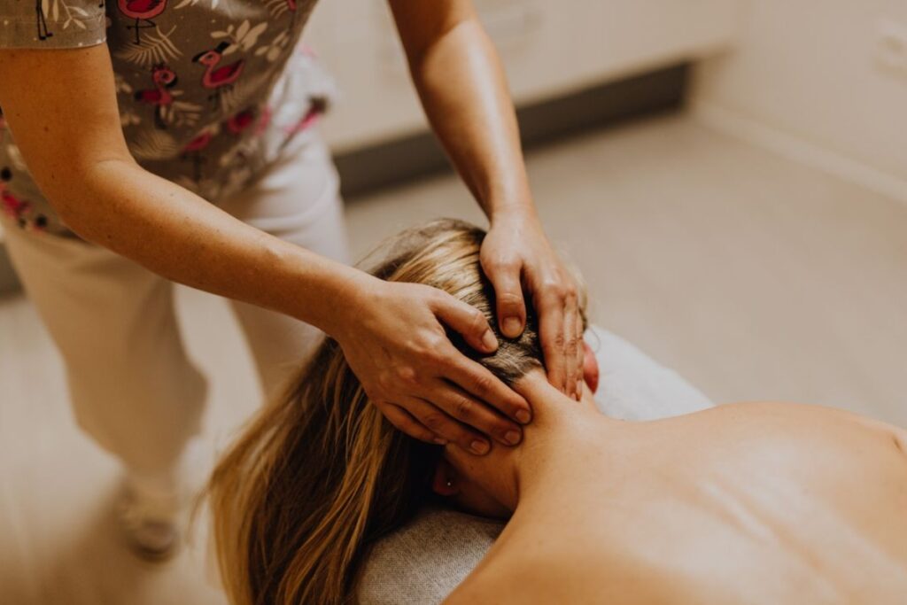 Fotografia em plano detalhe e ângulo superior mostrando as mãos de uma terapeuta pressionando e massageando a nuca e a base do crânio de uma mulher loira deitada de bruços em uma maca de massagem, em um ambiente clínico iluminado.